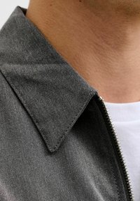 Primer plano del cuello de una chaqueta gris con costuras visibles y una cremallera plateada sobre una camisa blanca en el cuello y la parte inferior del rostro de una persona.
