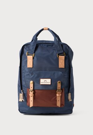 Navy blue backpack with brown bottom pocket, leather accents, gold hardware, and adjustable straps. Features a logo patch on the front.