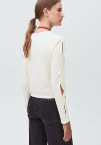 Ženska z dolgimi lasmi v čopku nosi belo dolge rokave s razporki na rokavih in črne hlače z okrasnimi žebljički v pasu.