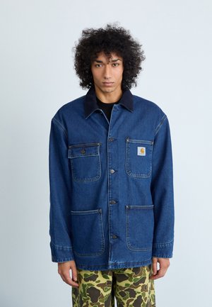 Denim jakna v temno modri barvi, z ovratnikom, dvema žepoma na prsih in logotipom. Obuta je čez kamuflažne hlače ohlapnega kroja.