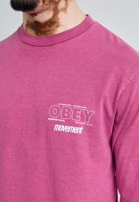 Camiseta de manga larga en algodón de color rosa brillante, con el texto blanco "OBEY PERPETUAL PERSISTENT movement" en el frontal, cuello redondo y corte relajado.