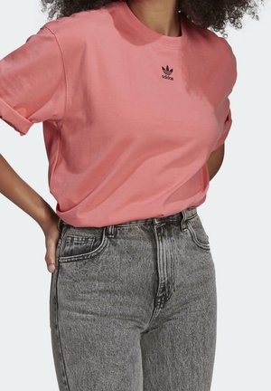 Mujer con camiseta rosa de Adidas con logo y vaqueros grises de cintura alta, de pie con una mano en la cadera.