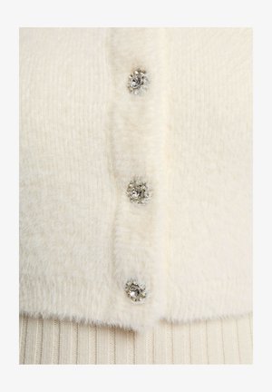 Cremefarbener, flauschiger Cardigan mit drei dekorativen Knöpfen, die mit klaren Strasssteinen versehen sind. Der Stoff hat ein weiches, strukturiertes Aussehen.