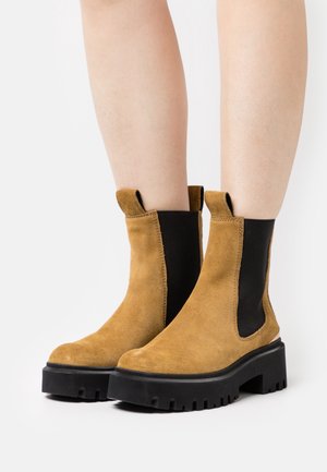 Botas de tobillo de ante marrón con una suela de goma negra gruesa, paneles elásticos en los laterales y lengüetas para facilitar su uso. Diseño de tacón plano.