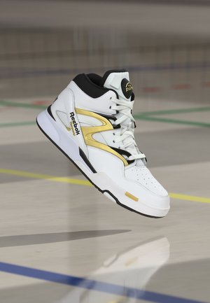 Biało-złoty wysokokrotny sneaker Reebok zawieszony w powietrzu na hali sportowej z widocznymi kolorowymi liniami granicznymi.