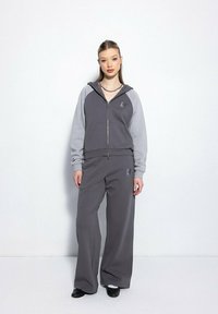 Młoda kobieta stoi przy białej ścianie, ubrana w dwukolorowy, szary sweatshirt na zamek i pasujące szerokie spodnie, a do tego czarne buty.