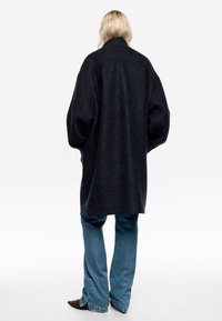 Pārmērīgi liels tumši pelēks vilnas mētelis ar augstu apkakli, nolaižamām plecu šuvēm un taisnu piegriezumu. Savienots ar gaiši zilām platām biksēm un tumšām kurpēm.