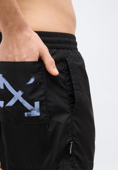 Gros plan d'une personne portant un short noir avec une taille élastique, la main dans la poche arrière présentant un motif de flèches croisées bleues.