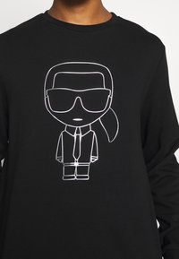 Černé tričko s bílou liniovou kresbou osoby, která nosí velké sluneční brýle, kravatu a culík v minimalistickém stylu.