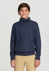 Granatowy sweter dzianinowy z wysokim kołnierzem w stylu golfowym, wzorem w warkocze oraz ściągaczami przy mankietach i dolnej krawędzi, zestawiony z beżowymi spodniami.