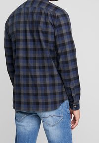 Hombre con una camisa de mangas largas a cuadros en azul, negro y gris, y unos vaqueros claros desgastados, visto desde la espalda contra un fondo liso.