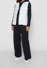 Donna che indossa un gilet imbottito azzurro chiaro sopra un felpa blu navy, pantaloni larghi blu navy e sneakers bianche, in piedi contro uno sfondo bianco.