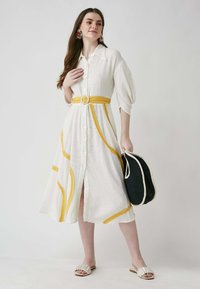 Weißes Hemdkleid mit gelben Akzenten und einem Gürtel, aus strukturiertem Stoff, mit kurzen puffärmel und knielangem Design; kombiniert mit einer schwarzen Tasche.
