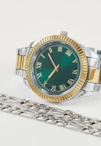 Montre en or et argent avec un cadran vert, des chiffres romains et une lunette texturée ; dotée d'un bracelet en chaîne métallique.