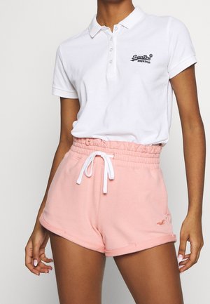 Kobieta w białej koszulce polo SuperDry i różowych szortach z wysokim stanem i troczkami, stojąca z rozluźnionymi rękami na jednolitym tle.