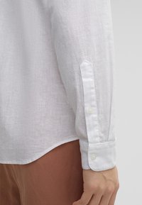Camicia bianca in lino a maniche lunghe con polsini abbottonati e orlo arrotondato, che mostra un tessuto testurizzato. Abbinata a pantaloni di tessuto marrone.