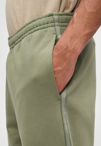 Ελιόχρωμο παντελόνι φόρμας με υφή, ελαστική μέση, πλαϊνές τσέπες και λευκή ετικέτα Lacoste στη ραφή.