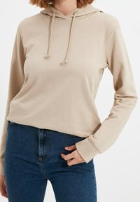 Sudadera beige con capucha y cordones, de mangas largas y corte relajado, combinada con unos pantalones vaqueros de denim azul de cintura alta. Textura suave visible.