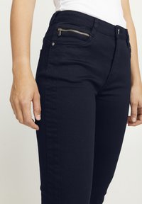 Jeans negros de cintura alta con un bolsillo con cremallera, ajuste elegante, textura suave y detalles en la costura lateral. Diseño clásico adecuado para un look casual.