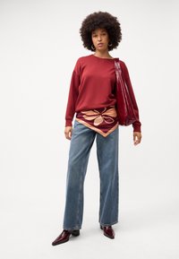 Felpa rossa, sciarpa con motivo annodata sotto, jeans larghi blu, stivali alla caviglia bordeaux lucidi e borsa a spalla bordeaux.