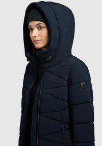 Chaqueta acolchada de color azul marino con una gran capucha, que presenta un diseño acolchado, cierre de cremallera y un parche con un logotipo redondo en la manga.