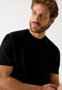 Sort kortærmet t-shirt lavet af blød bomuld, med et subtilt mønster af logoer i tonale nuancer. Rund halsudskæring og afslappet pasform.
