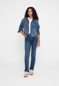 Chaqueta de mezclilla con cierres de botón, dos bolsillos en el pecho y mangas remangadas, combinada con jeans azul ajustados y zapatillas beige.