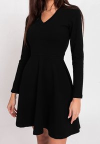 Robe noire à manches longues avec un col en V, un corsage ajusté et une jupe évasée. Confectionnée dans un tissu lisse et extensible, présentant un design simple.
