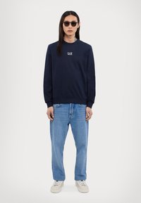 Jūras spēku lielais krekls ar logo, garām piedurknēm un ribotām aprocēm. Apvienots ar gaiši ziliem vaļīgiem džinsiem un krēmkrāsas kedām, modelis valkā saulesbrilles.