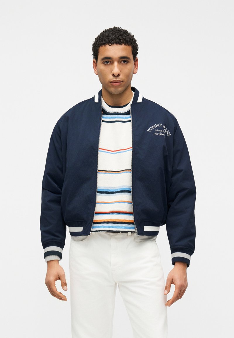 Άνδρας που φοράει μπλε ναυτικό μπουφάν τύπου μπόμπερ με λευκό λογότυπο, πολύχρωμο ριγέ πουλόβερ και λευκό παντελόνι, στέκεται μπροστά σε απλό φωτεινό φόντο.