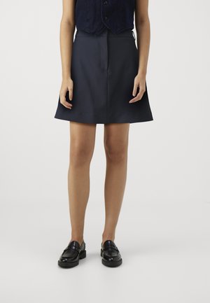 Woman wearing a navy blue skirt with a front seam, black loafers, and a dark sleeveless top, standing against a plain light background.