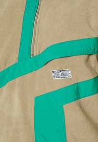 Sudadera de forro polar en color beige con detalles verdes. Cuenta con un cierre de cremallera de cuarto y un pequeño parche con el logo. Textura suave con paneles en contraste.