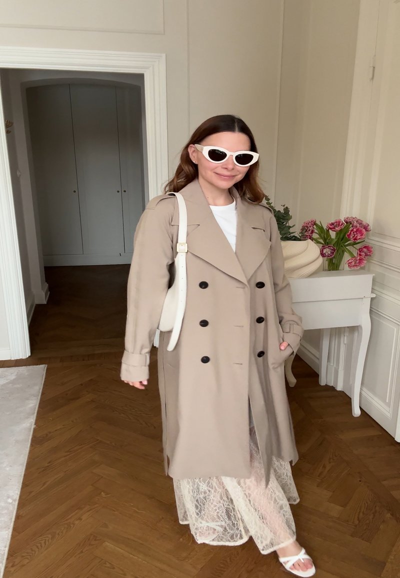 Femme portant un trench-coat beige, une jupe en dentelle blanche, des sandales blanches, des lunettes de soleil blanches et un sac à épaule, debout à l'intérieur près d'une table avec des fleurs.