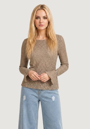 Donna bionda che indossa un top in pizzo color taupe a maniche lunghe con polsini svasati e jeans larghi azzurri segnati, in piedi davanti a uno sfondo semplice.