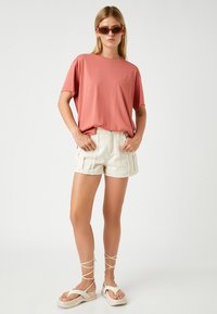Kortærmet koralfarvet bomulds-t-shirt i kombination med højtaljede cremede shorts og lyse strop sandaler på en lys baggrund.