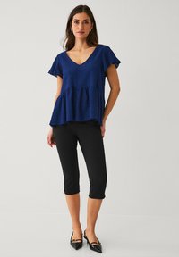 Marineblaues, strukturiertes Oberteil mit kurzen Flutter-Ärmeln, V-Ausschnitt und Peplum-Saum, kombiniert mit schwarzen, cropped Hosen und spitzen Pumps.