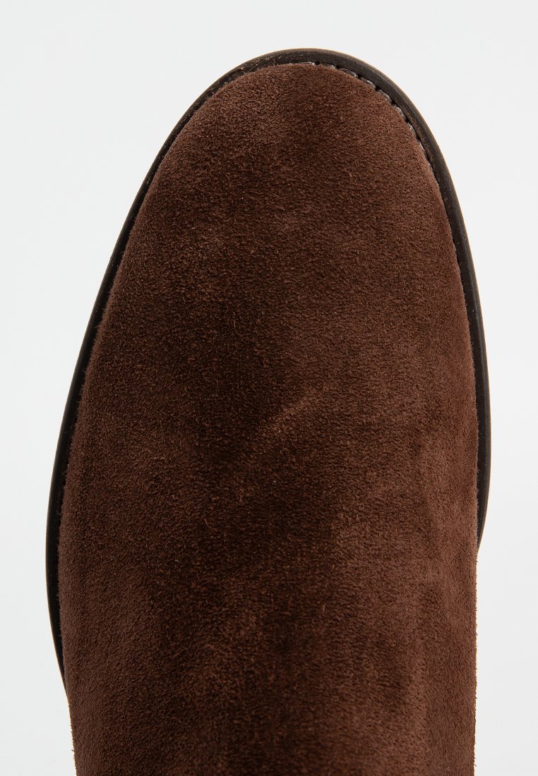 Brązowy zamszowy botek do kostki o gładkiej fakturze, zaokrąglonym nosku i czarnej krawędzi podeszwy. Skup się na miękkim wykończeniu górnej powierzchni.