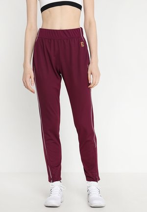 Kobieta ubrana w bordowe sportowe spodnie z białymi paskami po bokach i białe trampki, stojąca na tle jednolitego białego tła.