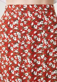 Jupe fleurie rouge avec des fleurs blanches et des feuilles vertes, fabriquée en tissu léger avec une texture lisse et une taille ajustée.