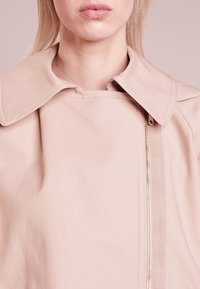 Gros plan d'une personne portant une veste beige avec un large col et une fermeture éclair asymétrique sur le côté droit.
