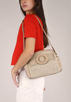 Bolsa estructurada beige con textura trenzada, detalle redondo con cuerda, tachuelas decorativas y una correa larga, llevada al hombro.