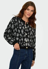 Femme portant un cardigan léopard noir et gris avec des boutons dorés, associé à un jean foncé taille haute, posant devant un fond uni.