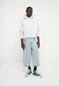 Γκρι φούτερ με κουκούλα και λογότυπο μπροστά, συνδυασμένο με ανοιχτό μπλε φαρδιά τζιν σόρτς και πράσινα ψηλά αθλητικά παπούτσια. Λευκό φόντο.