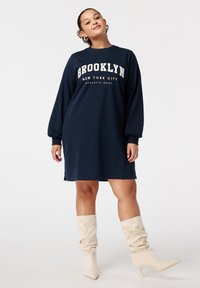 Robe sweat à manches oversize de couleur marine avec le texte "Brooklyn" et "New York City" en blanc. Associée à des bottes slouch beige clair montantes jusqu'aux genoux.