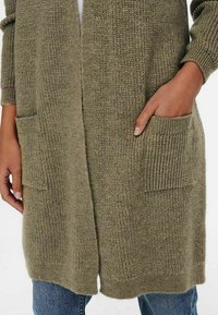 Cardigan lungo verde oliva con un motivo di maglia testurizzata, dotato di due tasche frontali e polsini a coste, indossato sopra jeans blu.