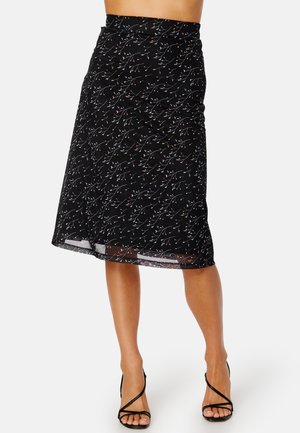 Jupe midi noire avec petits motifs floraux blancs et orange, portée avec des sandales noires à talons hauts et à brides, montrée de la taille aux pieds.