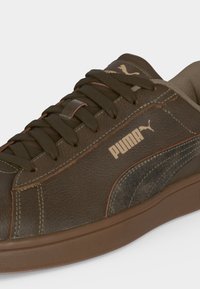 Baskets Puma marron avec lacets marron foncé, logo Puma beige et panneau latéral texturé sur un fond uni clair.