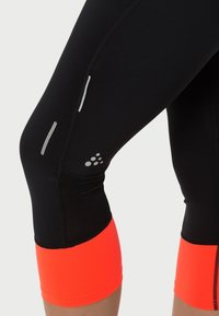 Leggings atléticos negros con una textura suave, con acentos en naranja neón en los puños. Incluye detalles reflectantes en los lados.