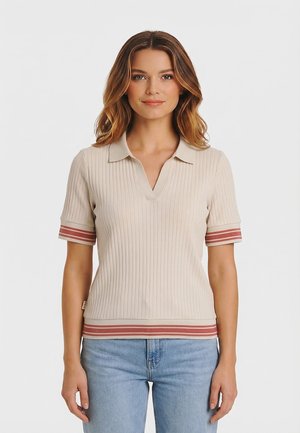 Mujer con cabello ondulado que lleva una camiseta polo de manga corta beige con canalé, con puños y dobladillo con rayas rojas, combinada con vaqueros azul claro.