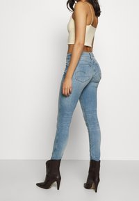 Mujer con jeans ajustados de color azul claro, una camiseta de tirantes recortada beige y botines negros de tacón, de pie de lado contra un fondo blanco.